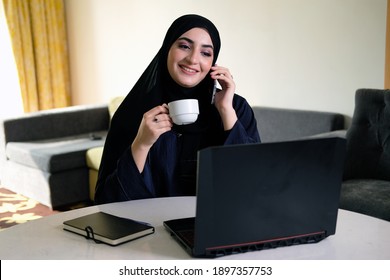 Multitasking Arabian Emirati woman wearing Shayla Abaya and Hijab using smart mobile phone and laptop while holding a coffee cup