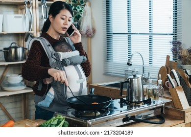 multitasking asian housewife carrying baby with a carrier is speaking on the smartphone while cooking breakfast using frying pan in the kitchen at home. - Powered by Shutterstock