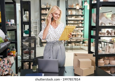 Multitasking. Cheerful Mature Business Woman, Shop Owner Talking On The Phone, Holding Envelope While Checking Her Mail, Standing In Craft Pottery Shop