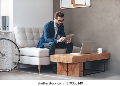 Multitasking. Handsome young man wearing glasses and working with touchpad while sitting on the couch in office - Shutterstock ID 370353149