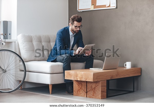 Multitasking. Handsome\
young man wearing glasses and working with touchpad while sitting\
on the couch in\
office