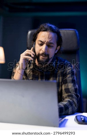 Multitasking individual efficiently manages a conversation on his mobile device while simultaneously working on digital laptop. Male person having remote communication with clients on cell phone.