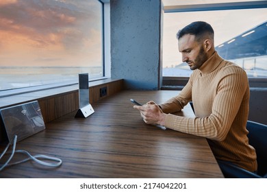 Multitasking man browsing the internet on his smartphone, sitting by windows overlooking runway and beautiful sky at sunset in the VIP lounge of an international airport, waiting to board the flight - Shutterstock ID 2174042201