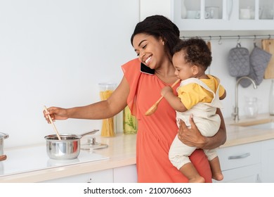 Multitasking Mom. Smiling Black Woman Holding Baby, Cooking And Talking On Cellphone In Kitchen Interior, Happy African American Mother Stirring Food In Pot And Taking Care About Toddler Son