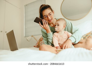 Multitasking Mother Taking A Phone Call While Sitting On A Bed With Her Baby. Single Mom Making Plans With Her Clients While Working From Home. New Mom Balancing Motherhood And Work.