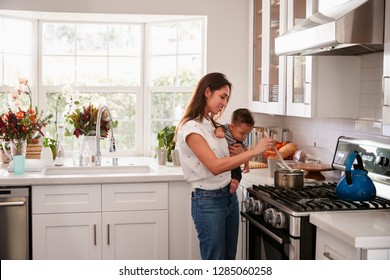 Multitasking Mum Holding Her Young Baby While She Makes Food At The Hob In Her Kitchen, Side View