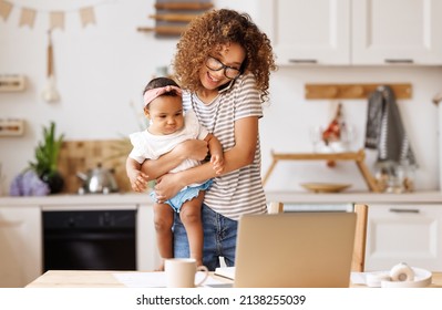 Multitasking Young African American Woman, Working Mother Freelancer  Using Smartphone And  Laptop Computer With Baby Girl In Hands, Ethnic Female Doing Freelance Work On Maternity Leave