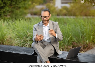 Multitasking young businessman in park relaxing with smartphone and coffee, smiling male entrepreneur in suit sitting on bench with laptop beside him, embodying modern work flexibility - Powered by Shutterstock