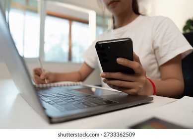 Multitasking. Young individual using smartphone while taking notes on laptop.