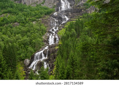 Multi-tier waterfall streaming down rocky cliff amid lush green forest in Norway, summer landscape with flowing water and natural scenery.