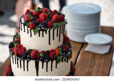 Multi-tier Wedding Cake On A Wooden Table With Berries On Top And Chocolate Running Down The Sides Of The Cake With Two Stacks Of Plates In The Background.