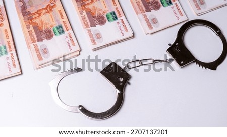 A multitude of five thousand Russian banknotes, arranged in a fan-like formation, rests on a wooden table, against a backdrop of black police handcuffs. The idea of economic wrongdoing, larceny, or