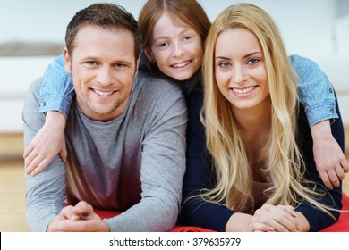 Mum, Dad And Their Cute Young Daughter Relaxing Together On A Rug On The Floor Facing Towards The Camera With Happy Friendly Smiles