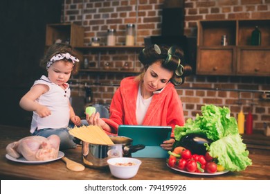 Mum at home. Young mother with little child in the home kitchen. Woman doing many tasks while looks after her baby. Use a tablet PC. Technology and Maternity.