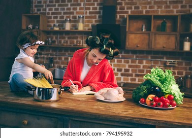 Mum at home. Young mother with little child in the home kitchen. Woman doing many tasks while looks after her baby. She talking on a cell phone or smartphone, writing a pen to do list in a notebook.