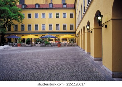 Munich, Bavaria, Germany - July 1, 2009: Summer view arch pillars with light and plaza against tables with parasols at an open-air café of a yellow building
