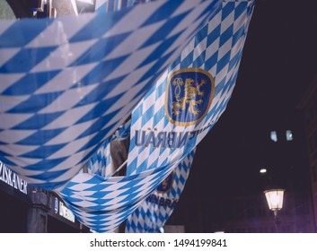 Munich, Germany - Aug. 31, 2019: Löwenbräu Brewery Bavarian Blue White Flag Hanging At House In Munich Night Dark Evening Time Illuminated