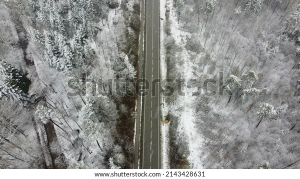Munich, Germany\
Bavaria - April 4, 2022: Striped paved winter road winding through\
the snowy forest trees with a bike path and cars. Image taken from\
a drone at 100m\
altitude.