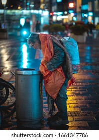 MUNICH, GERMANY - Feb 01, 2020: Homeless Man Looking For Drinks And Food In Garbage Bin. Cold Rainy Night In Munich With Wet Streets Reflecting The Lights Of The City