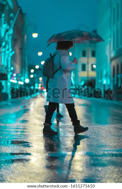 MUNICH, GERMANY - Feb 01,\
2020: People crossing the street during a wet rainy night in\
Munich. Neon lights illuminating the scene. City traffic in the\
background.