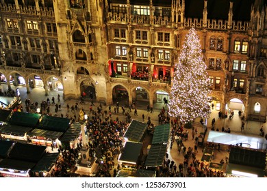 Munich Old City Square Aerial View With Huge Christmas Tree, Market Stalls And Lights. Illuminated Shops, City Council At Marienplatz With People And Tourists Drinking Mulled Wine And Winter Shopping