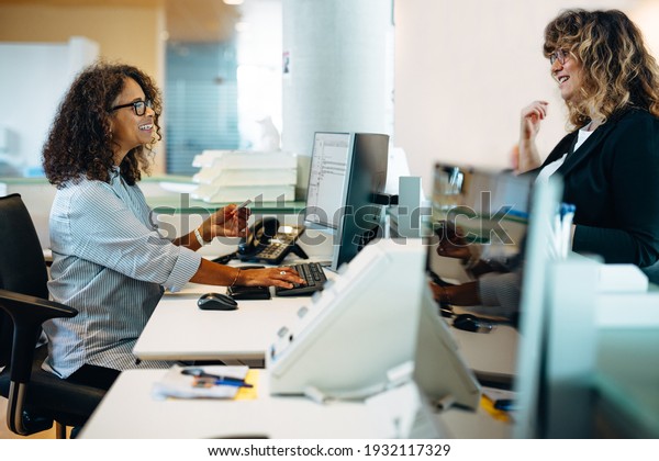 Municipal office administrator assisting a woman\
standing at her desk. Friendly receptionist helping a woman\
visiting municipality\
office.