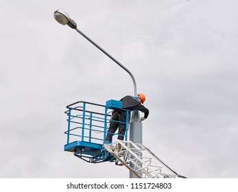 Municipal Worker Man With Helmet And Safety Protective Equipment Painting Street Lightning Pole At Height With Brush. Worker Repair Light Pole. High Elevated Cherry Picker With Worker On Floodlight