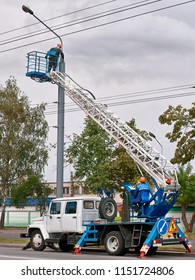 Municipal Worker Man With Helmet And Safety Protective Equipment Painting Street Lightning Pole At Height With Brush Tool. Worker In Lift Bucket Repair Street Light Pole. Technician On Aerial Device