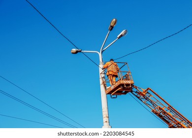 A Municipal Worker In Protective Equipment Performs Hazardous Work To Eliminate An Interruption In The Power Grid. A Worker Repairs A Street Lamp From An Aerial Platform Basket.