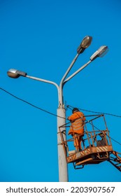 A Municipal Worker In Protective Equipment Performs Hazardous Work To Eliminate An Interruption In The Power Grid. A Worker Repairs A Street Lamp From An Aerial Platform Basket.