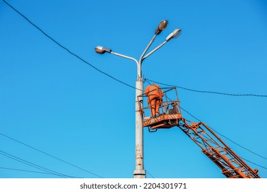 A Municipal Worker In Protective Equipment Performs Hazardous Work To Eliminate An Interruption In The Power Grid. A Worker Repairs A Street Lamp From An Aerial Platform Basket.