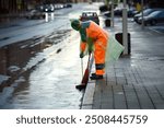 Municipal worker in raincoat cleanup street with broom and dustpan, street workers keep city clean in all weather conditions. Man with broomstick sweeping road, collect garbage in scoop in the evening