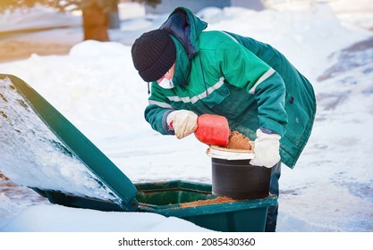 Municipal worker in uniform scoop sand from grit bin, spreading deicing chemicals on slippery sidewalk. Sanding street, prevent injury and slipping accident