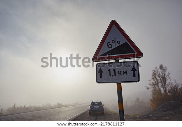 Murmansk region, Russia -
September 2017:  Road sign a steep descent of 6 percent, amid a fog
gray sky. Nissan Terrano car on a background of bright foggy sky
with the sun
