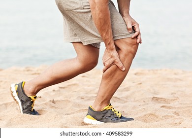 Muscle injury. Man with sprain thigh muscles. Athlete in sports shorts clutching his thigh muscles after pulling or straining them while jogging on the beach. - Shutterstock ID 409583299
