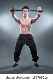 Muscled Asian Kung Fu Man In Action Pose With Nunchucks. Blood Stripes On His Chest. Wearing Black Pants. Studio Shot Against Grey. 