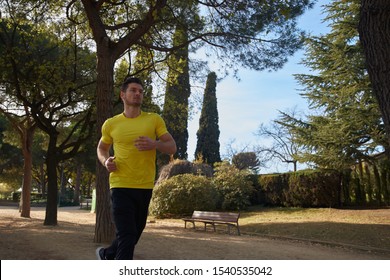 Muscled Young Man In A Yellow Shirt And Black Sweat Pants Runs Through A Park On A Sunny Day As He Exercises