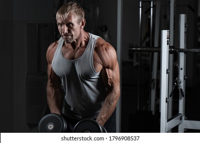 Muscular adult brutal man with dumbbells pumps biceps in the gym. Portrait of caucasian authentic bodybuilder doing workout exercises