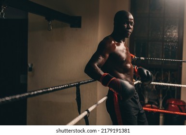 Muscular African boxer looking at the camera while resting in his corner of the boxing ring. Athletic young man having a training session in a boxing gym.
