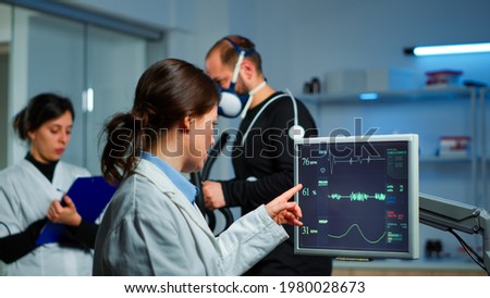 Muscular athlete with mask running on cross trainer in medical laboratory for monitoring his performance while ekg scan runs on computer screen and team of doctor researcher examining results