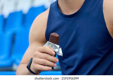 Muscular Athlete Snacks On A Protein Bar While Resting After A Workout At The Stadium. Man In A Blue Sports Jersey Rests In The Stand For Fans, Eating A Chocolate Bar.