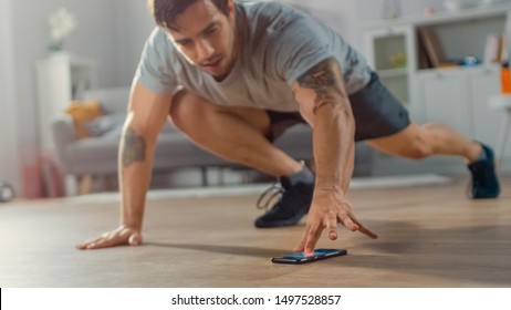 Muscular Athletic Fit Man in T-shirt and Shorts is Doing Mountain Climber Exercises While Using a Stopwatch on His Phone. He is Training at Home in His Bright Living Room with Minimalistic Interior.