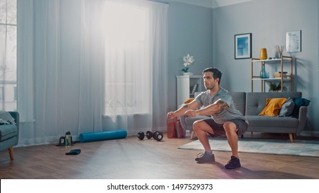 Muscular Athletic Fit Man in T-shirt and Shorts is Doing Squat Exercises at Home in His Spacious and Bright Living Room with Minimalistic Interior. - Shutterstock ID 1497529373