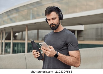 Muscular athletic man looks at smartphone screen downloads songs in playlist for training holds bottle with fresh water wears casual t shirt has regular workout poses outdoors. Healthy lifestyle - Powered by Shutterstock