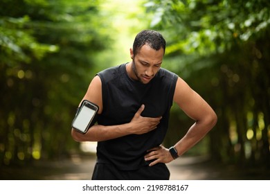 Muscular Attractive Black Man With Heart Pain Touching His Chest, Jogging In The Public Park, Using, Armband With Smartphone, Mockup, Copy Space. Sport And Cardiovascular Diseases Concept