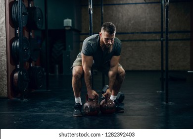 Muscular Attractive Caucasian Bearded Man Lifting Two Kettlebells In A Gym. Weight Plates, Dumbbell And Tires In Background.