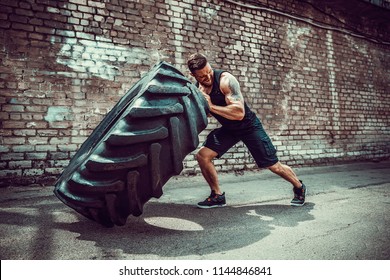 Muscular bearded tattooed fitness man moving large tire in street gym. Concept lifting, workout training.