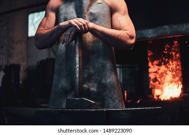 Muscular blacksmith in protective apron at his workshop with hammer in his hands. There are fire at background. - Powered by Shutterstock