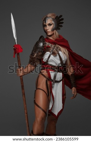 A muscular blonde Viking woman in fantasy armor with a red cloak, baring her thighs, poses holding a spear and shield against a gray background