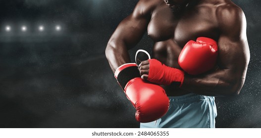 A muscular boxer adjusts his red boxing gloves in a dimly lit boxing ring, ready for a fight. - Powered by Shutterstock